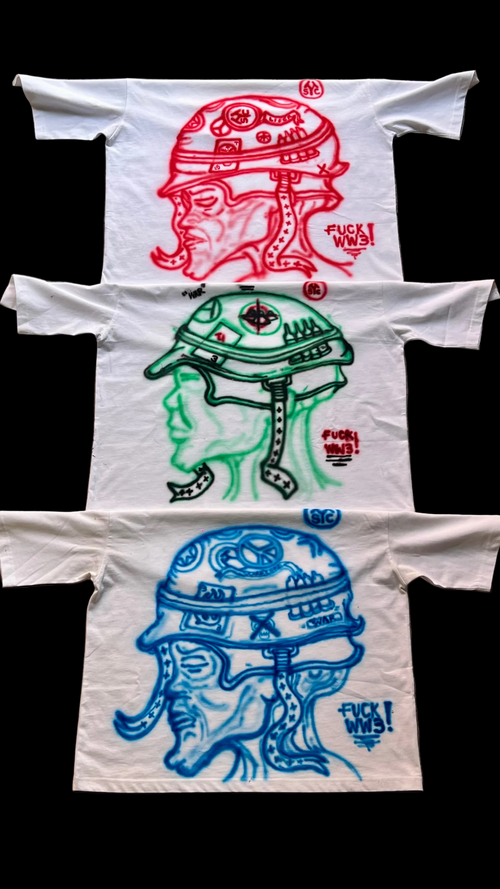 Three white graphic T-shirts laid flat on a black background, each featuring a stylized helmeted face illustration in a different color: red at the top, green in the middle, and blue at the bottom. Each shirt also includes small red text near the right chest area.